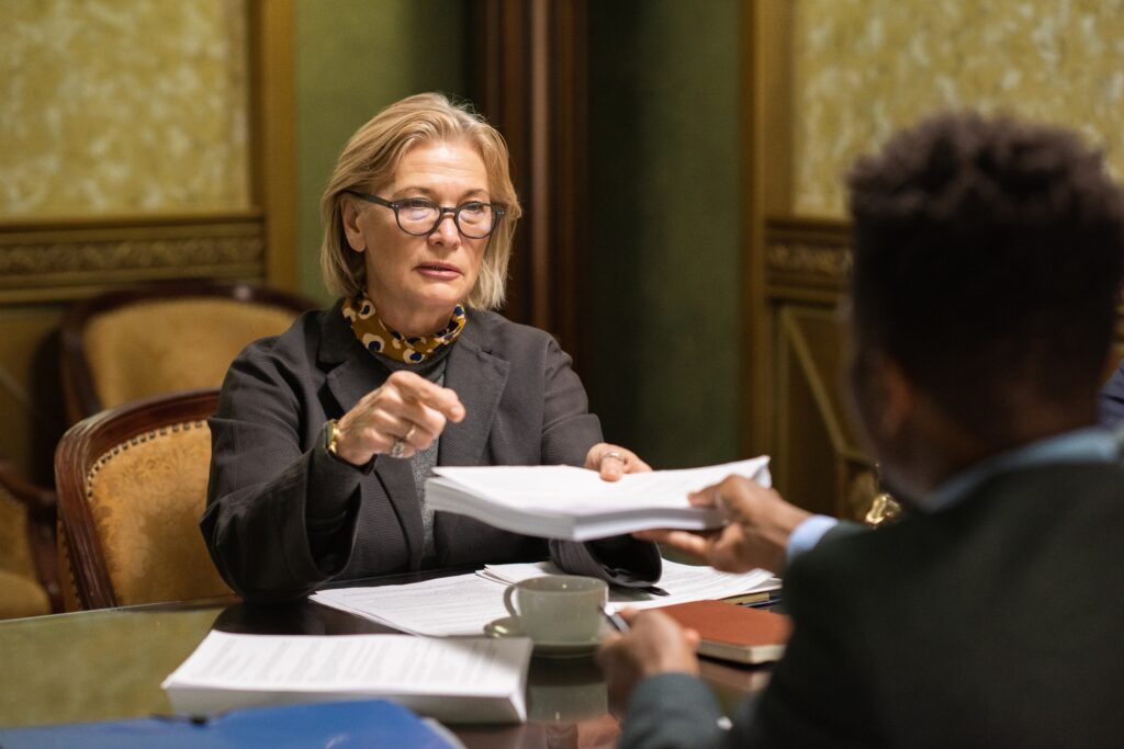 An older woman sits in a business meeting in a luxurious office. She is surrounded by papers.