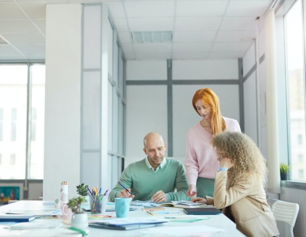 A team of three coworkers sits in a brightly lit office around a table covered in papers and coffee mugs. There is a white man and two women, one white and one of an unidentified race.