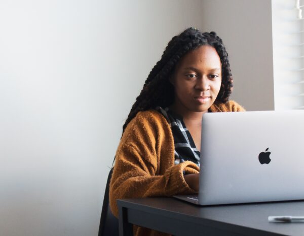 A black woman sits at a table with a laptop. She has long braided hair and wears a yellow sweater.