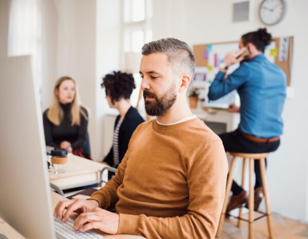 A man sits at a computer in a busy office setting. He wears a brown sweater.