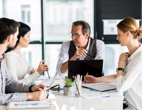 A group of people sit around a table in a brightly lit office. There is one older man, two younger women, and one younger man. They wear professional clothing.