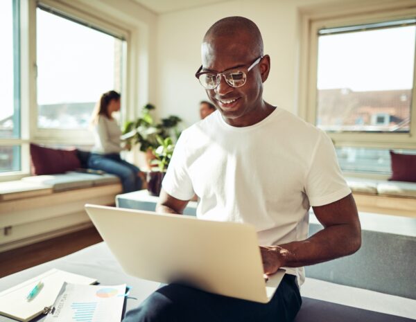 A man sits with a laptop in a brightly lit office setting. He has a shaved head and wears a white tshirt and glasses.