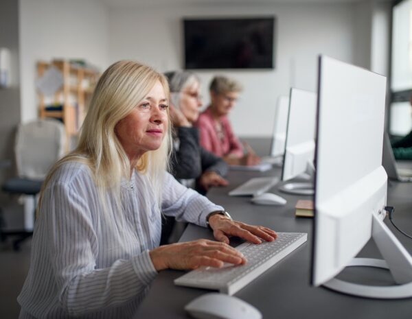 An older woman sits at a desktop computer in a modern office setting. She has light blonde hair and wears a white shirt.