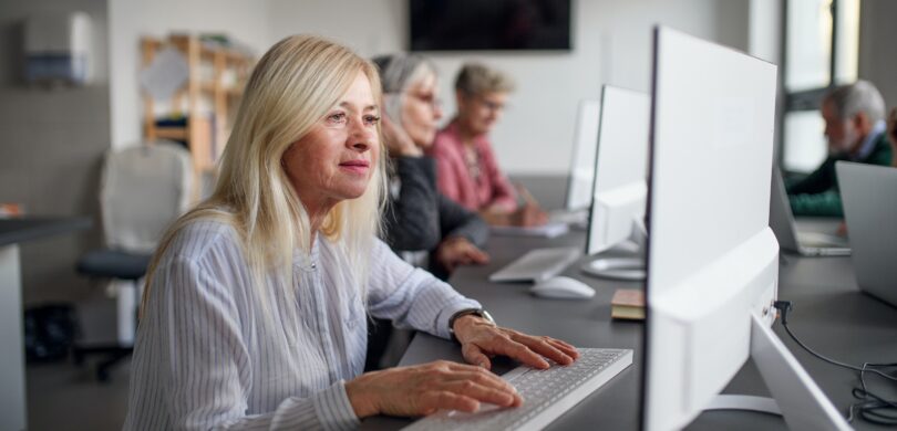 An older woman sits at a desktop computer in a modern office setting. She has light blonde hair and wears a white shirt.