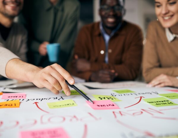 A team of diverse professionals sits in the background. They are looking at a large piece of paper covered in writing and colorful post-it notes.