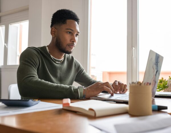 A black man sits at a desk in front of a computer and pads of paper. He wears a dark green long-sleeved shirt and a beaded necklace. He has short, dark hair.